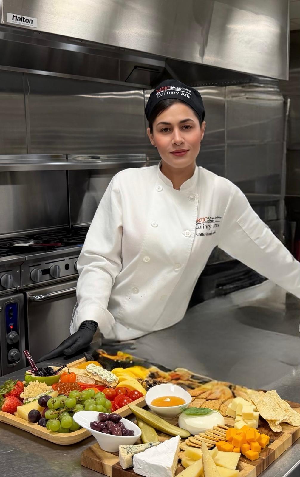 Chef Cinthia in her chef's whites standing behind a grazing board of fruit, cheeses, and crackers