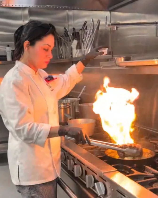 Chef Cinthia in chef whites, flaming a pan on a professional range